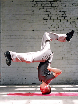 DR BEAT in a headstand freeze with no hands on floor and legs kicked out. white brick background with concrete floor and red lino. Press shot for Freshmess tour of "COMBO/TABLES". taken in DanceBase's studio 2 during construction.